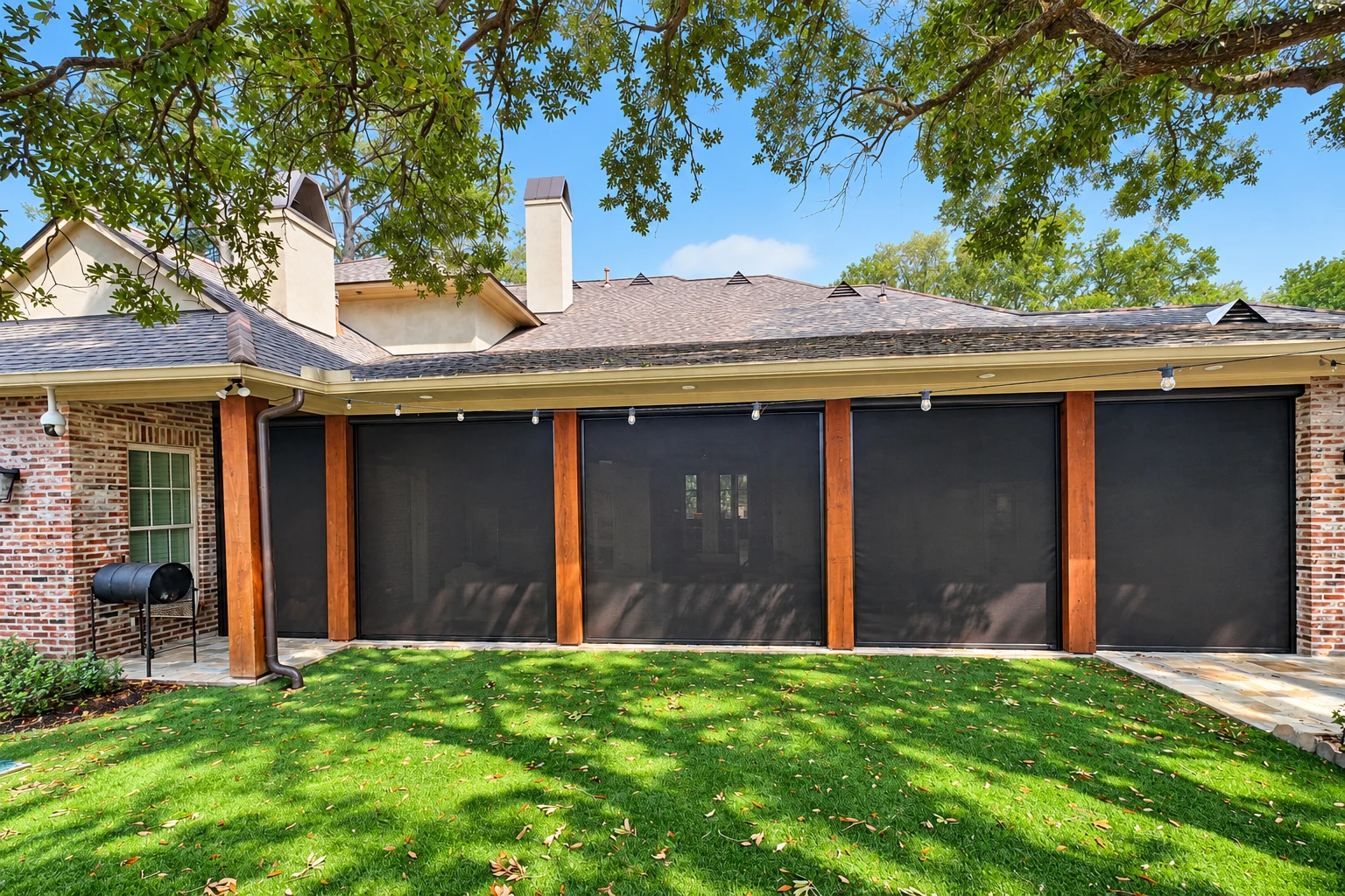 Enclosed patio with dark screen panels and wood posts beneath a broad roof, opening onto a green lawn.