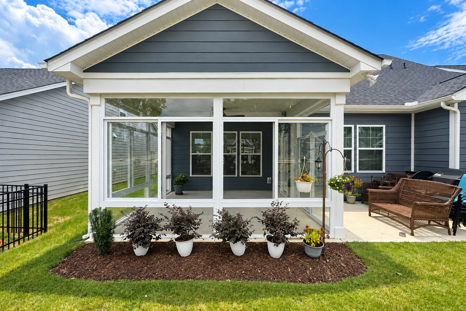 A glass-enclosed patio room with white framing stands beside a blue-gray house, with potted plants along the front.