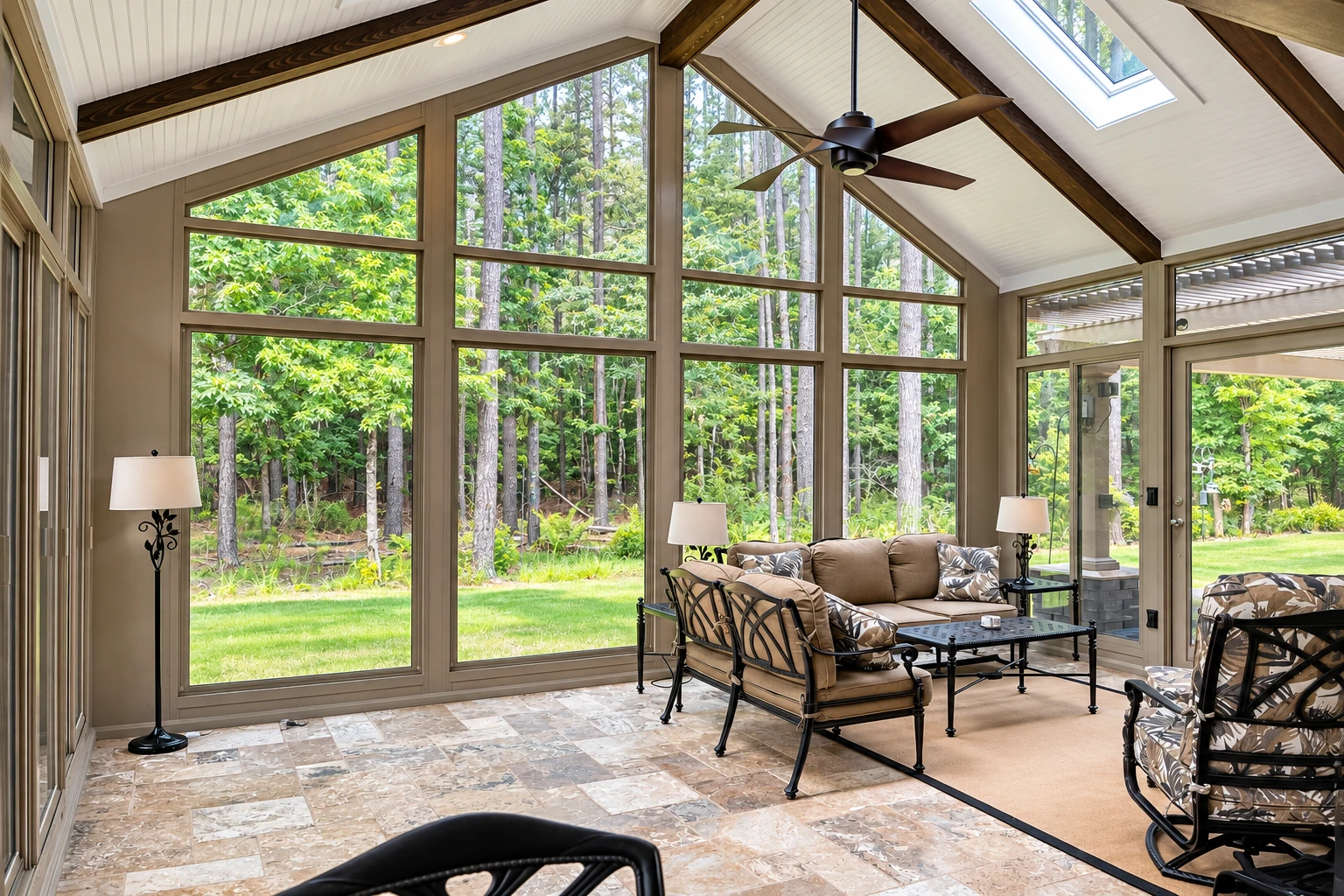 A sunroom addition with a vaulted white ceiling, exposed dark beams, tall windows overlooking trees, and a seating area.