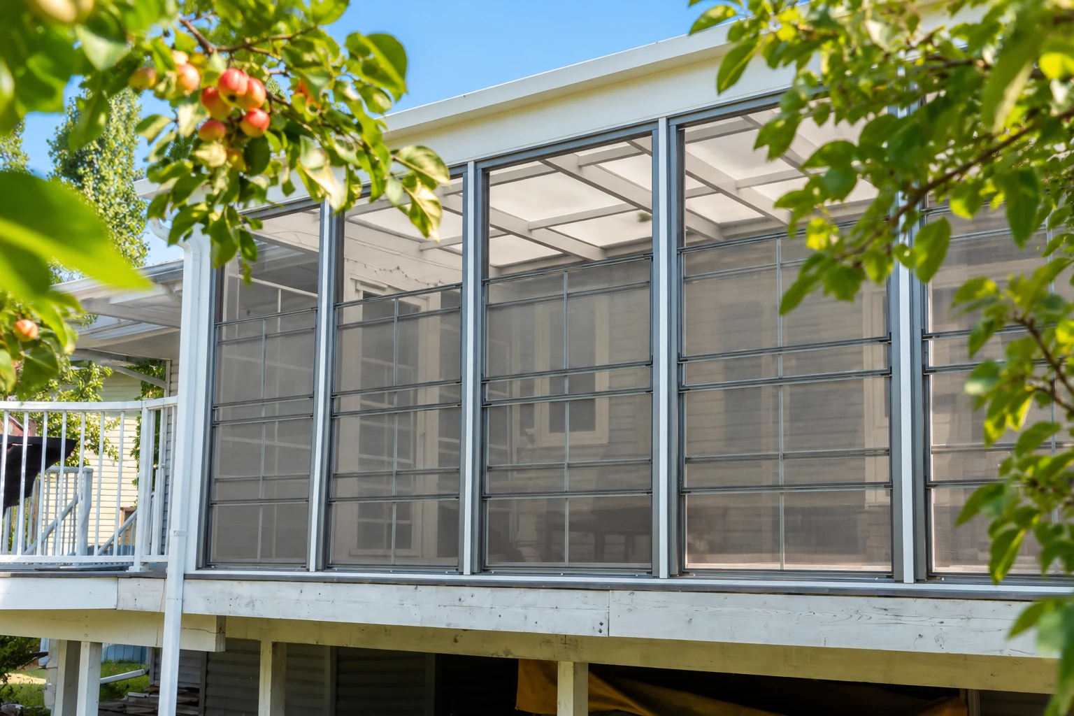 Screened sunroom under construction on a raised deck, with dark framed panels and leafy branches in the foreground.