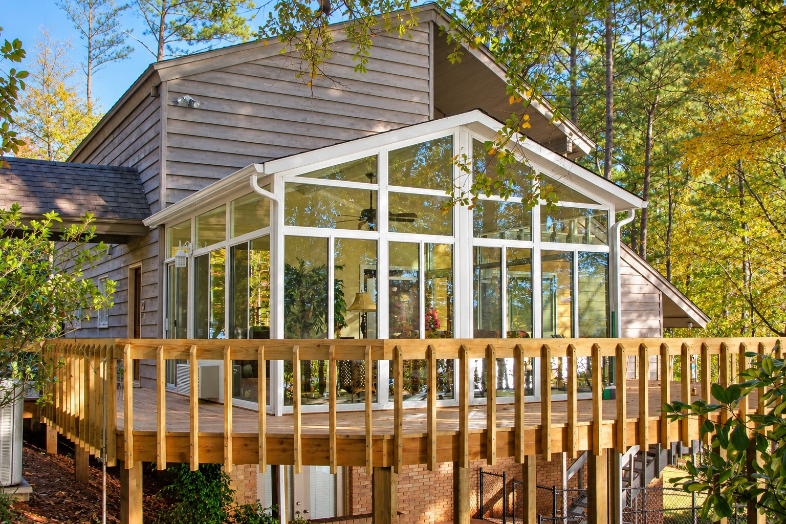 A white, gable-roofed vinyl sunroom with extensive windows sits on a raised wooden deck among trees.